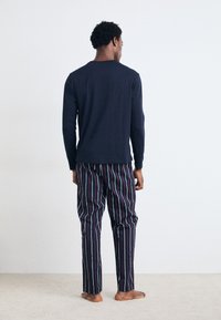 Marineblå langærmet skjorte parret med flerfarvede stribede bukser, der har en afslappet pasform og blød tekstur. Fødderne er bare mod et lyst tæppe.