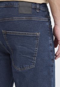 Tumši zili džinsa džinsi ar teksturētu apdari, kam ir melnas ādas jostasvietas etiķete un kontrastējošas šuvju detaļas.