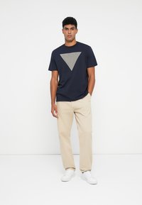 Camiseta de marino con un gráfico triangular gris en el pecho. Combinada con pantalones beige claros y zapatillas blancas.