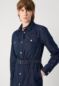 Denim dress in dark blue with a fitted silhouette, button front, two chest pockets, and a belted waist. Long sleeves with button cuffs.