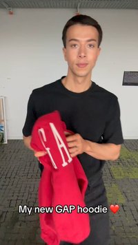 Νέος άνδρας με μαύρο πουκάμισο κρατάει μια κόκκινη μπλούζα με κουκούλα GAP σε εσωτερικό χώρο με το κείμενο "Η νέα μου μπλούζα GAP" και ένα κόκκινο emoji καρδιάς.