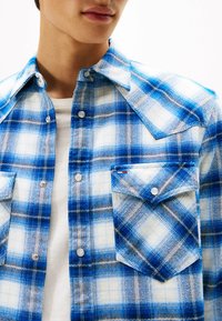 Blauw-witte geruite flanellen shirt met drukknoopjes, spitse kraag en twee borstzakken. Beschikt over een gestructureerde stof in een ruitpatroon.