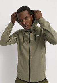 Olive green zip-up hoodie with a textured surface, adjustable drawstrings, and black accents along the zipper and seams.