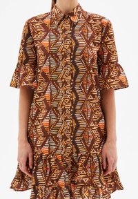 Abito camicia marrone con motivi geometrici e astratti in arancione, giallo e crema. Presenta maniche arricciate e una chiusura a bottoni sul davanti.