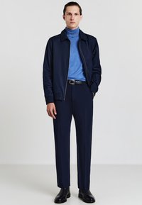 Marinefarbener Reißverschlussjacke über einem hellblauen Rollkragenpullover, kombiniert mit dunkelblauen Hosen und schwarzen Lederschuhen; glatte Materialien und maßgeschneiderte Passform.