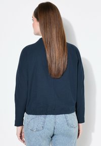 Giacca cropped navy con maniche lunghe, texture morbida e vestibilità rilassata, abbinata a jeans a vita alta blu chiaro con tasche posteriori.