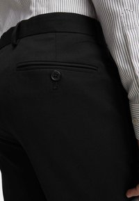 Czarne spodnie o gładkiej fakturze, z tylną kieszenią zapiętą na czarny guzik i subtelnymi detalami stębnowania. Częściowo widoczna koszula w paski.