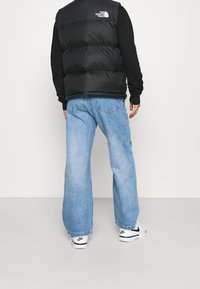 Gilet imbottito nero con collo alto, abbinato a jeans larghe a gamba larga di colore azzurro chiaro e scarpe sportive bianche con dettagli neri.