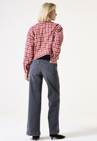 Vrouw die met haar rug naar ons toe staat, gekleed in een rood-wit geruite jas, wijde grijze jeans en zwarte lage hakken.