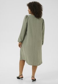 Robe verte avec des pois noirs, manches bouffantes longues, décolleté modeste et une silhouette en A à hauteur du genou. Portée avec des sandales à talons noires.