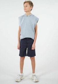 Hoodie sans manches gris avec cordon de serrage et logo, associé à un short bleu marine. Le modèle porte des baskets claires avec des lacets réfléchissants.