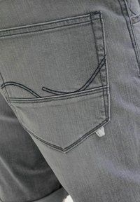 Grijze denim jeans met een achterzak met een gebogen naaddetail, subtiele textuur en een klein wit label nabij de rand van de zak.