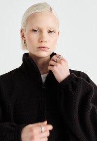 Schwarze Fleecejacke mit hohem Kragen und halbem Reißverschluss, die eine strukturierte Oberfläche und voluminöse Ärmel aufweist. Silberne Ringe an den Händen.