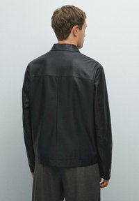 Man wearing a black leather jacket and gray trousers, facing away against a plain pale gray background.