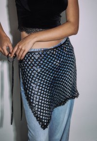 Crni mrežasti predložak s valovitim rubom, pričvršćen crnim vezicama, preko svijetloplavih traperica, s sjajnom, teksturiranom površinom.