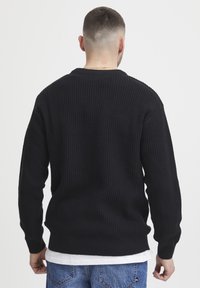Black ribbed sweater with long sleeves, crew neck, and textured fabric. Worn over a white shirt, paired with light blue jeans.
