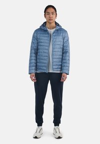 Giacca imbottita azzurro chiaro con cappuccio, chiusura con zip e polsini elasticizzati, abbinata a una maglietta grigia e pantaloni blu navy. Le sneaker bianche completano il look.
