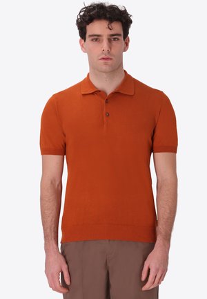 Uomo con capelli ricci che indossa una polo in maglia arancione a maniche corte e pantaloni marroni, in piedi davanti a uno sfondo chiaro uniforme.