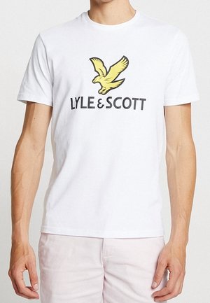 Mann trägt ein weißes T-Shirt mit gelbem Adler-Logo und dem schwarzen Schriftzug "LYLE & SCOTT" auf der Brust, kombiniert mit hellen Hosen.