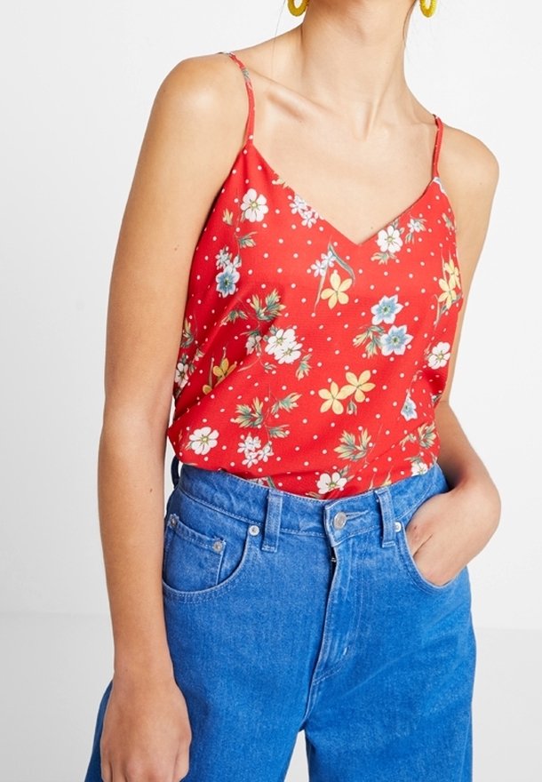 Femme portant un haut à bretelles fines rouge à motifs floraux, rentré dans un jean bleu taille haute, une main dans la poche, sur un fond blanc.