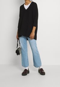 Czarny oversizeowy sweter z dzianiny z dekoltem w serek, zestawiony z jasnoniebieskimi dzwonami. Modelka nosi ciemne lakierowane loafersy i trzyma małą czarną torebkę.