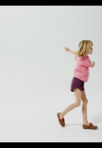 Young child wearing a pink shirt and dark shorts running or skipping on a plain white background.