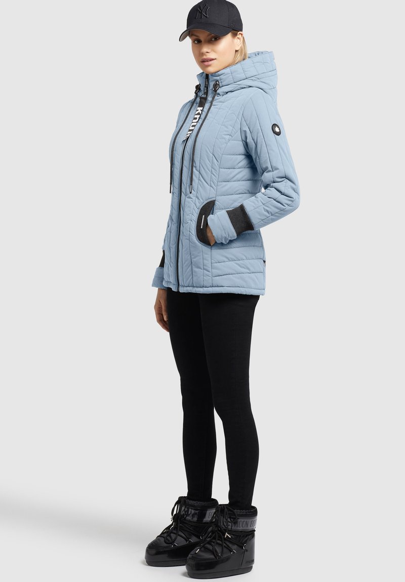 Frau in leichter blauer Steppjacke, schwarzen enganliegenden Hosen, schwarzen Winterstiefeln und schwarzem Baseballcap, leicht nach rechts gedreht.