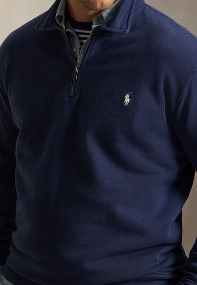 Άνδρας που φοράει σκούρο μπλε πουλόβερ με φερμουάρ, γκρι πουκάμισο με γιακά και ριγέ μπλούζα από κάτω, με μικρό κεντημένο λογότυπο.