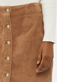 Brun corduroy skjørt med en teksturert overflate, med sølvknapp-tilbehør langs forsiden. Hånd synlig på siden.