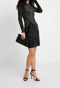 Svart långärmad klänning med en glittrig textur och diagonala sammetsdetaljer, tillsammans med en svart texturerad clutch och sandaler med remmar.