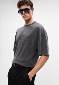 Graues, oversized T-Shirt mit kurzen Ärmeln, das vorne mit einem dezent geprägten "HUGO"-Logo versehen ist. Schwarze Hosen und Sonnenbrille runden den Look ab.