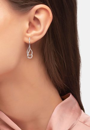 Pendiente en forma de gota de plata con pequeños cristales incrustados, llevado en la oreja izquierda, contrastando con el largo cabello castaño y el collar de color rosa pálido.