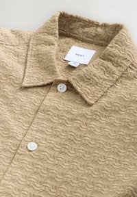 Chemise beige à texture avec un col, présentant un motif de feuilles, deux boutons blancs, et une étiquette portant la mention "NEXT" avec une taille "M".