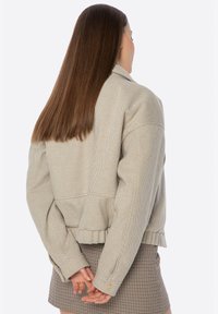 Giacca beige a righe con design corto, maniche larghe e orlo con dettaglio elastico. Modello mostrato di spalle con le mani intrecciate.