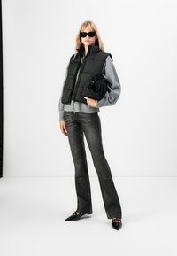 Vrouw in zwarte zonnebril, zwarte puffer vest over een grijze trui, zwarte leren broek, met een zwarte tas in de hand, staand tegen een witte achtergrond.