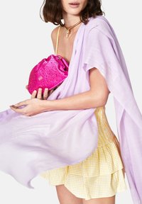 Borsa a sacco con paillettes fucsia e texture morbida, tenuta con una mano. Sciarpa trasparente lavanda sovrapposta a una mini gonna a quadretti gialli.