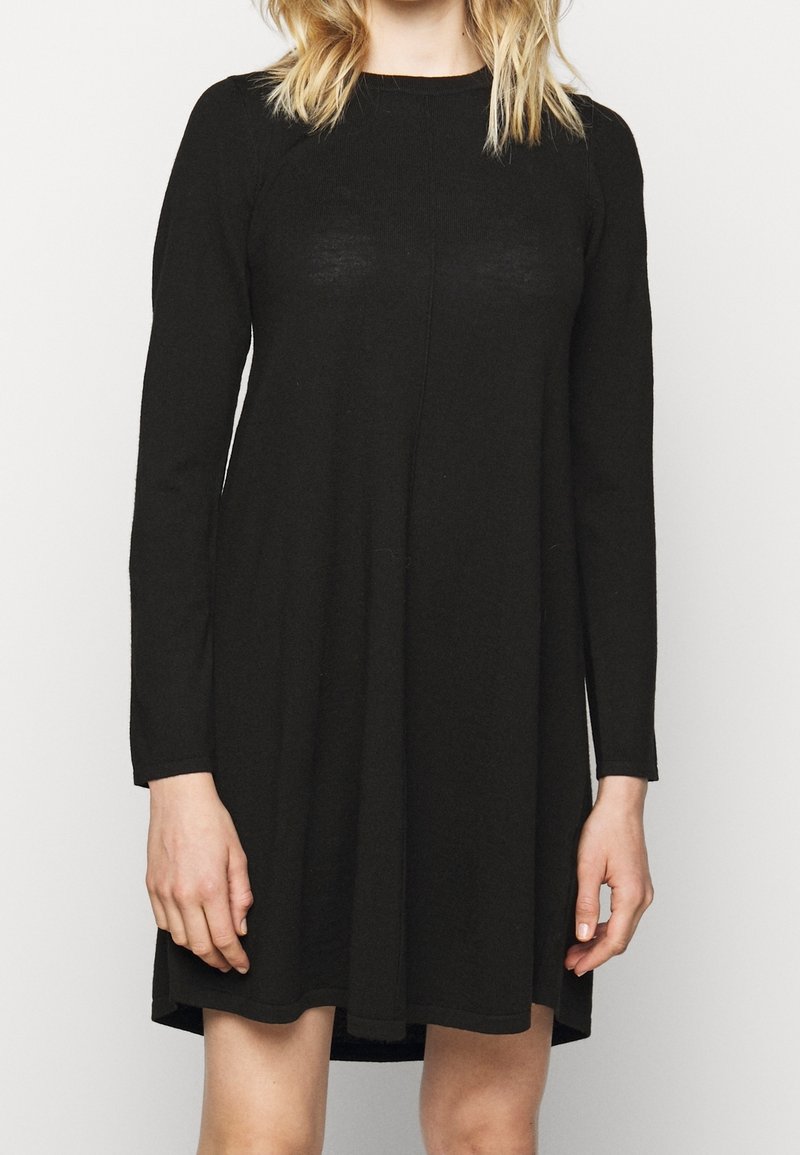 Femme portant une robe noire à manches longues, longueur genou, debout devant un fond blanc uni.