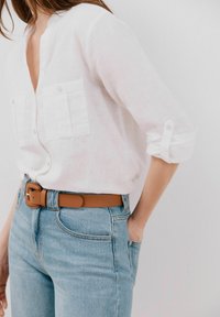 Blusa di lino bianca con scollatura a V, due tasche frontali, maniche arrotolate. Indossata con jeans a vita alta di colore blu chiaro e una cintura marrone.