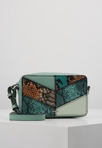 Piccola borsa a tracolla rettangolare con toppe geometriche multicolori in turchese, marrone e beige, con una tracolla removibile su una superficie bianca.