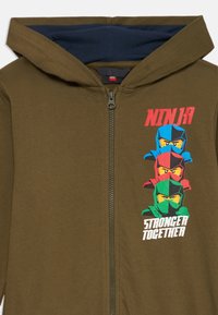 Sweatshirt verde-azeitona com fecho éclair e forro da capuz em azul-marinho. Apresenta uma imagem de três personagens ninja coloridos e o texto "NINJA MAIS FORTE JUNTOS."