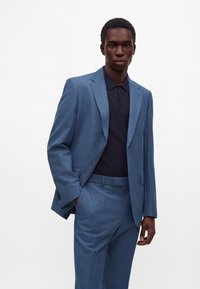 Jeune homme portant un costume bleu clair avec un pantalon assorti et une chemise boutonnée bleu marine foncé, debout devant un fond blanc uni.