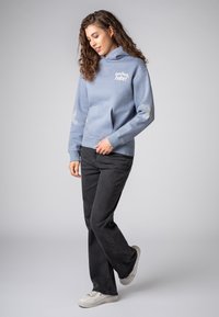 Lichtblauwe hoodie met een voorzak, voorzien van witte tekst en patroonaccenten op de mouwen. Combinerend met zwarte jeans en witte sneakers.