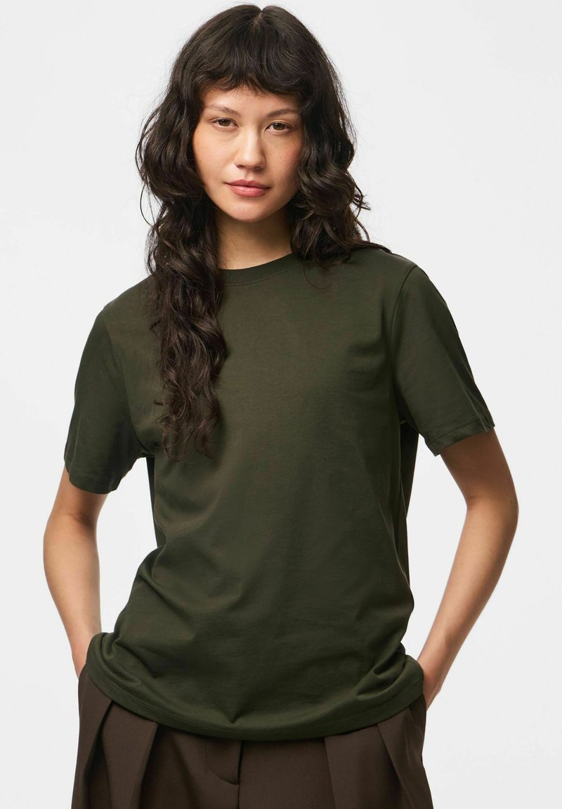 Jeune personne aux longs cheveux ondulés foncés, portant un t-shirt uni vert foncé à manches courtes et un pantalon marron, les mains dans les poches.