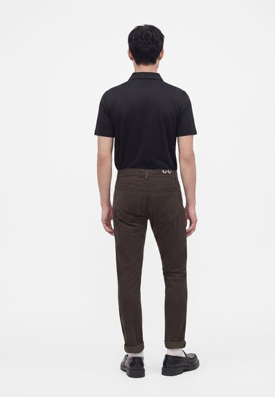 Homme debout de dos, portant un polo noir à manches courtes, un pantalon marron foncé avec des revers, des chaussettes blanches et des chaussures noires sur un fond blanc uni.