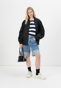 Casaco bomber preto, t-shirt riscada em branco e azul marinho, calções em denim, mala preta, sapatilhas brancas com detalhes em bege e meias pretas.