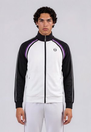 Un jeune homme est debout face à nous, portant une veste de sport blanche et noire zippée avec des rayures violettes et un pantalon blanc assorti.