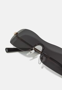 Gafas de sol negras con una montura ancha y curvada, que presenta un detalle dorado en el puente y almohadillas nasales transparentes, con patillas de acetato negro.