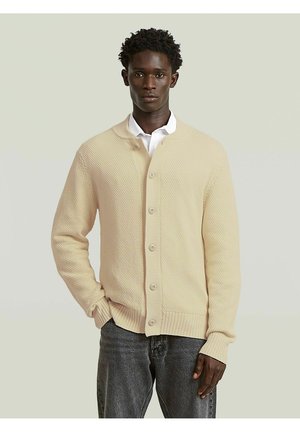 Man die een beige gebreide gebuttoniseerde cardigan draagt over een wit overhemd en donkere spijkerbroek, staand tegen een eenvoudige lichte achtergrond.
