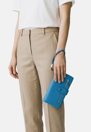 Persona che indossa pantaloni beige e una camicia bianca che tiene in mano un portafoglio da polso blu con texture, chiusura con cerniera e bottone a scatto.