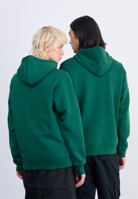 Des sweatshirts à capuche verts en tissu doux, présentant une coupe décontractée, des poignets et un ourlet côtelés, vus de dos sur deux personnes.